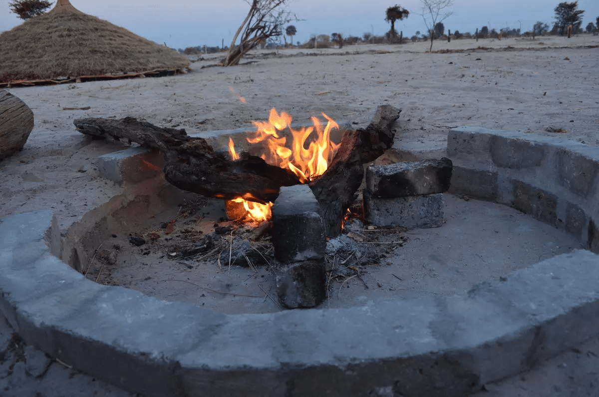 xongula-village-homestead-resdest-namibia-north-owamboland-campfire1.jpg.pagespeed.ic_.5xip1vkFJj a fire pit in the sand with a hut in the background
